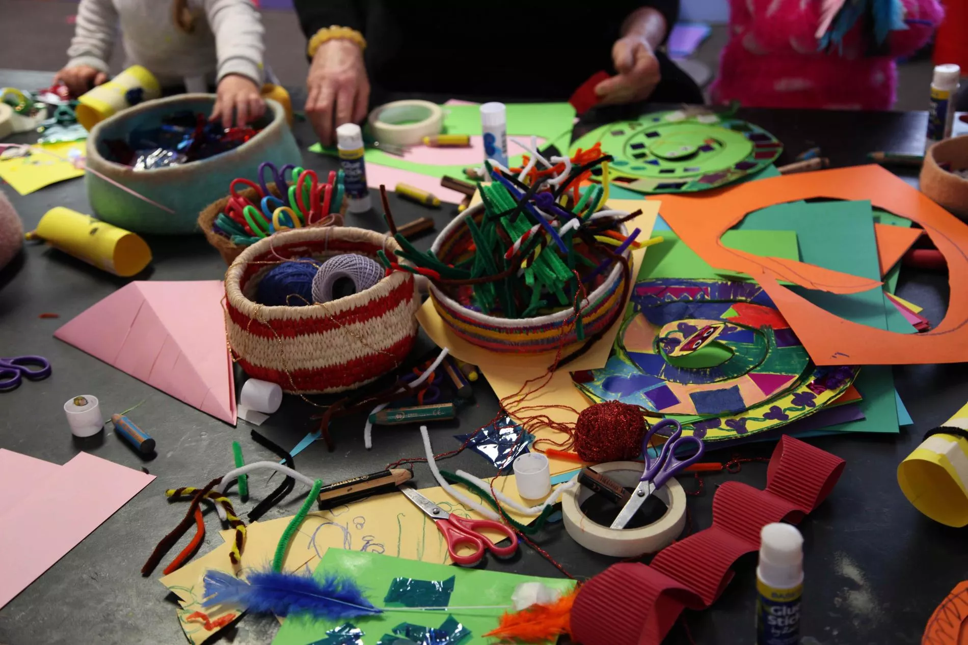 A table covered in coloured paper, feathers, scissors, glue, string and craft supplies. 