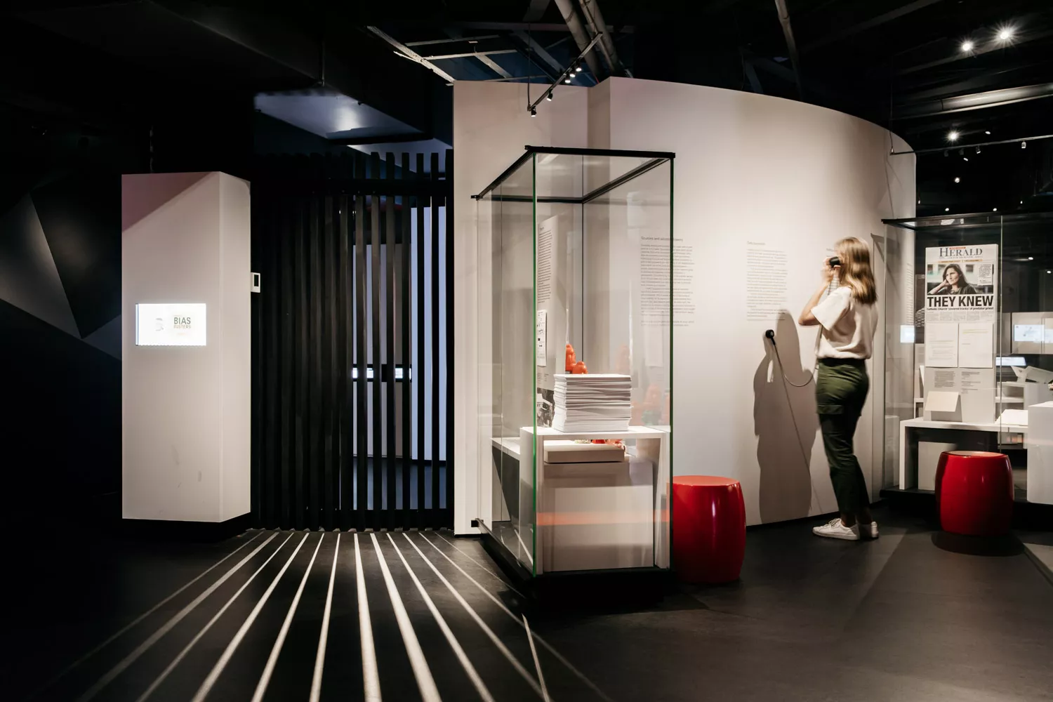 A woman listens to an audio device in an exhibition surrounded by glass panels with papers in them. 