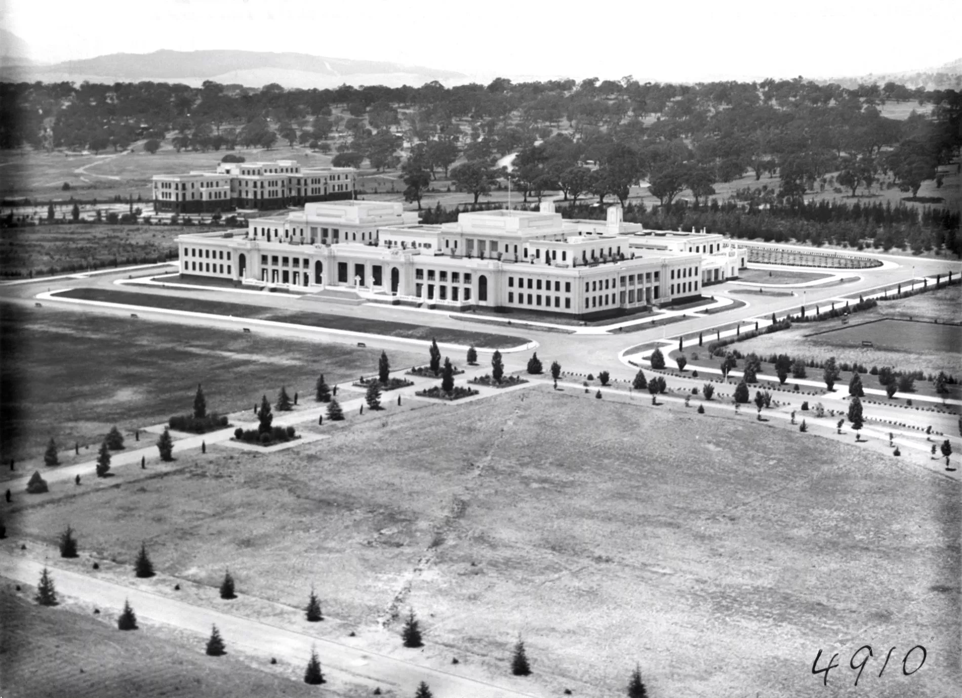 A black and white arial shot of a Old Parliament House, a large white building, surrounded by newly cleared land with planted trees. 