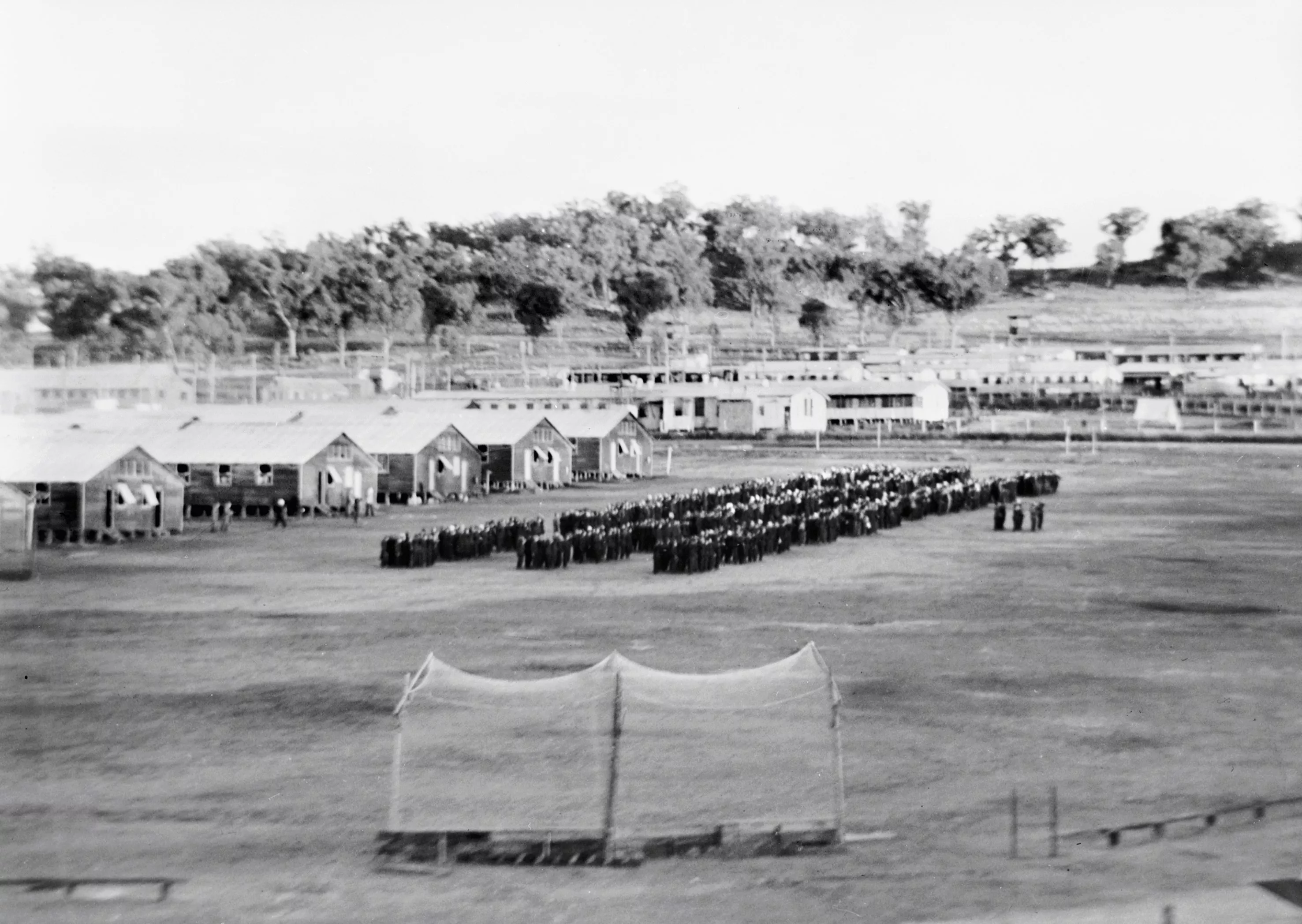 Lines of Japanese prisoners seen from far away, lined up in a ritualistic manner in front of many military huts inside an internment camp.     