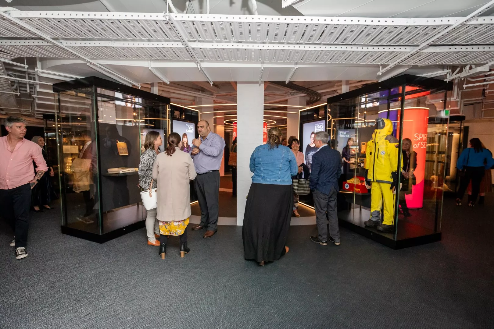 People mill around talking to each other and looking into glass displays in a museum exhibition. One features a bright yellow suit.