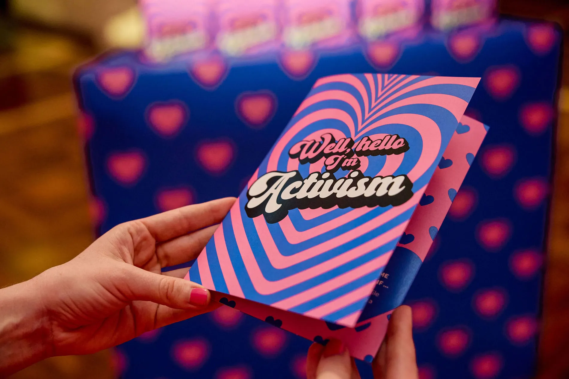 A pink and blue card with the text 'Well, hello I'm activism' is held by a person with pink nails.