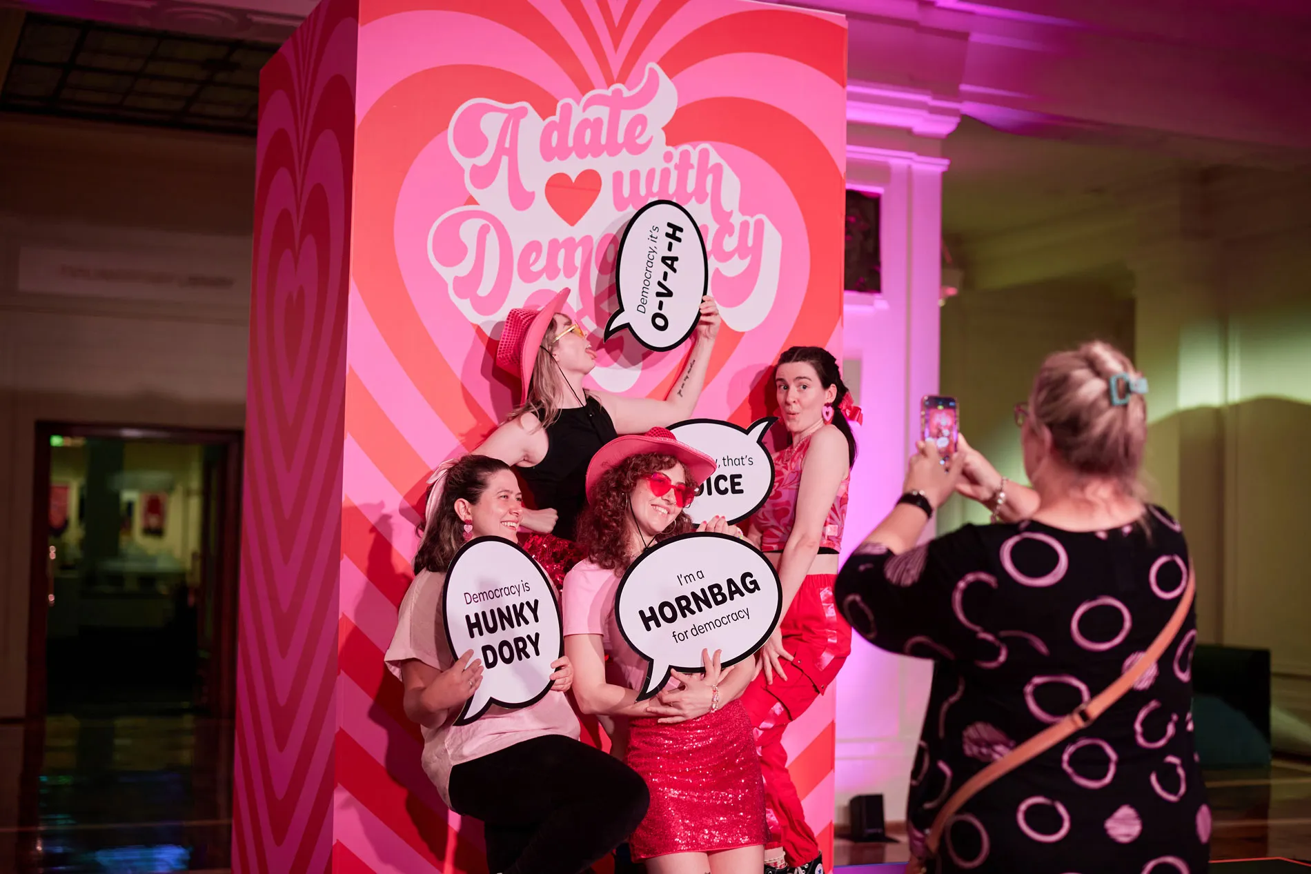 Four women hold signs in front of a pink and red backdrop with the text  'Date with democracy' in Old Parliament House.