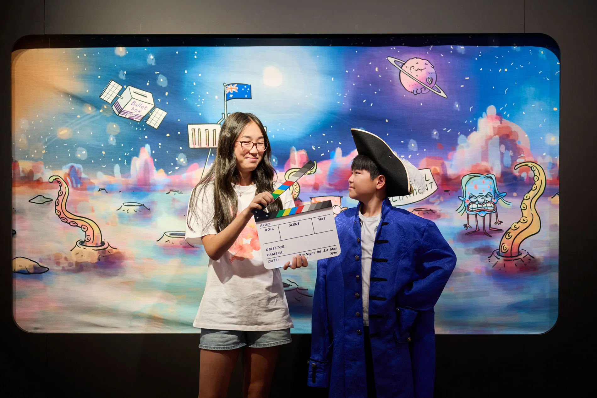 Two kids stand in front of a graphic cartoon backdrop, one holds a movie clapperboard to call 'action'.